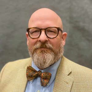 Chris MacDonald is a white man with a beard wearing a tan suit, blue shirt, bowtie, and glasses